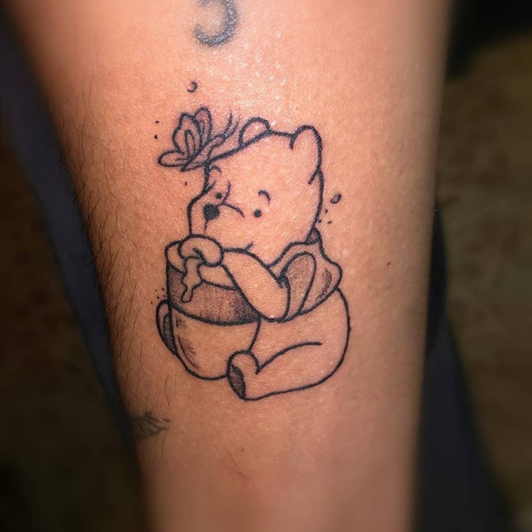 Outline tattoo of Winnie the Pooh sitting with a honey pot and a butterfly on his head on a person's leg.